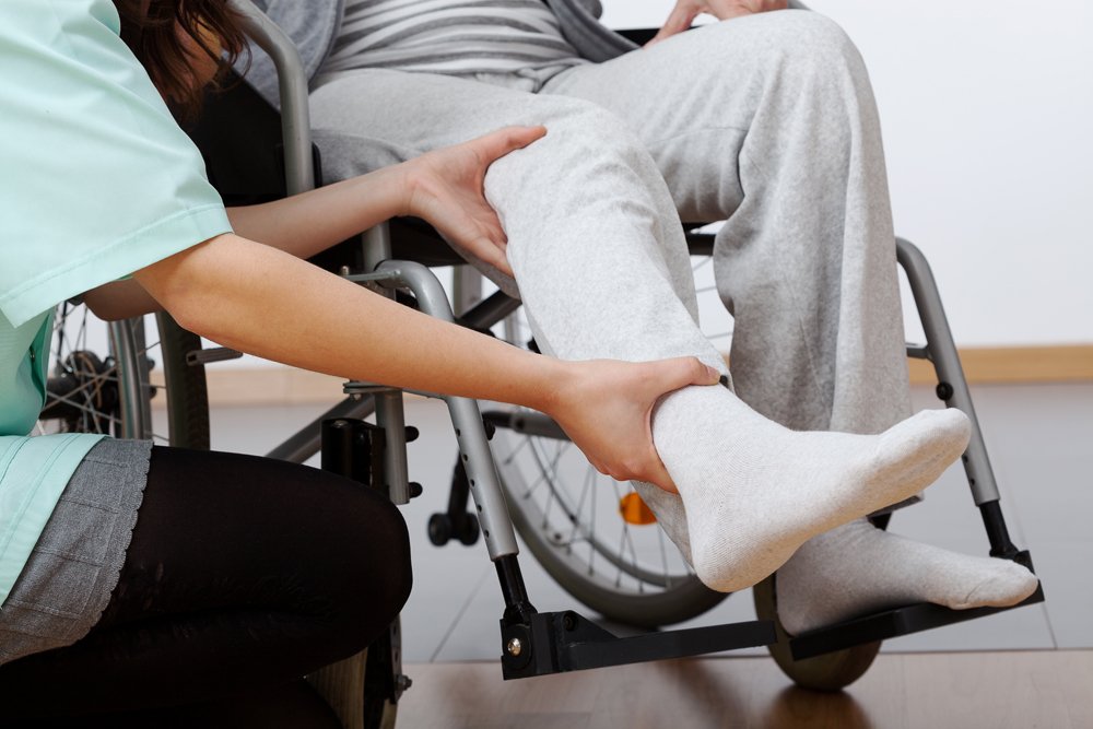 Descrição da imagem #pracegover: Uma pessoa está sentada na cadeira de rodas. Um profissional da área de saúde segura uma de suas pernas. Fim da descrição.