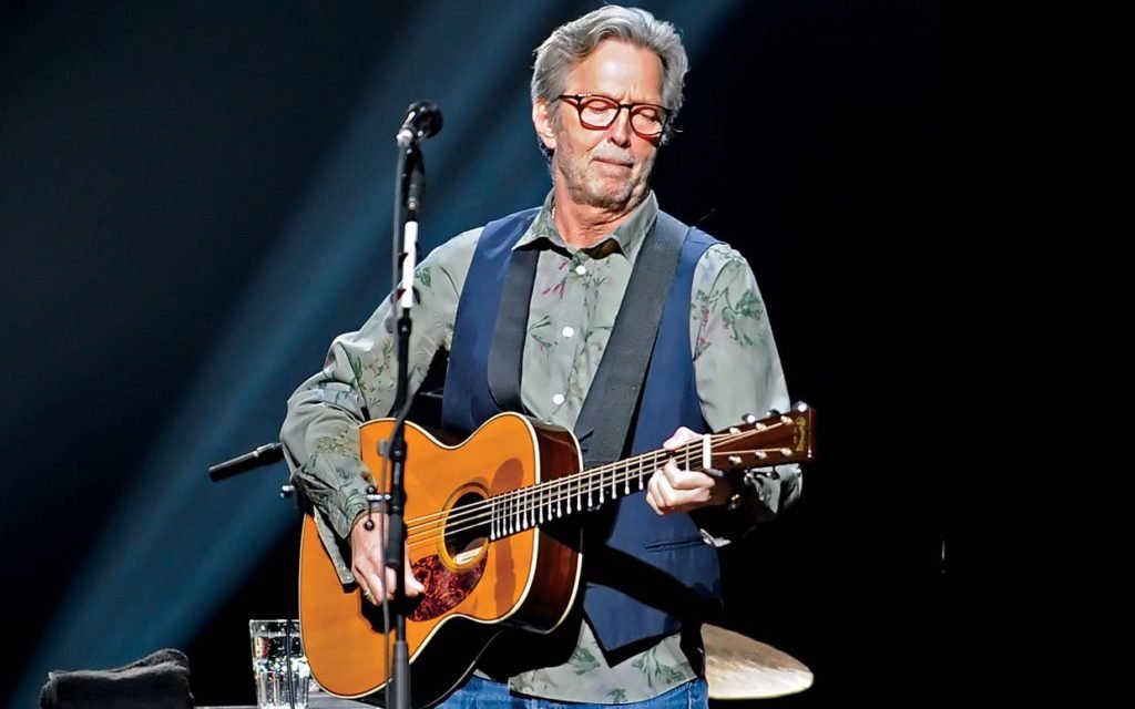Descrição da imagem #PraTodosVerem: Imagem no formato retangular, na horizontal. O músico Eric Clapton, durante um show. Ele está com um violão. Eric é um homem grisalho, de pele morena e cabelos lisos. Fim da descrição.