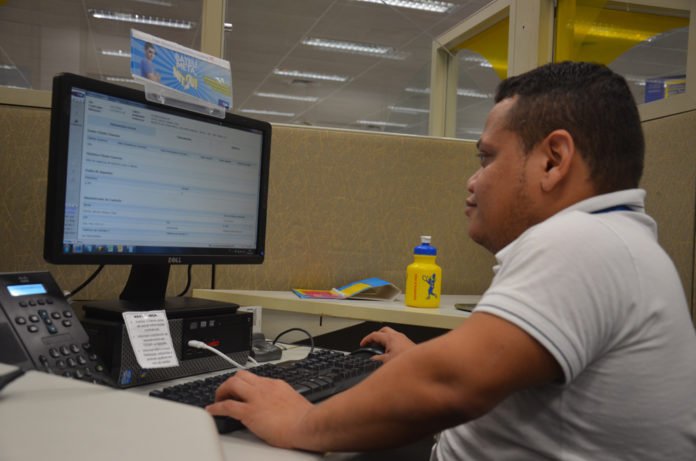 Descrição da imagem #PraTodosVerem: A imagem está no formato retangular, na horizontal. O colaborador José Nairton está sentado em sua estação de trabalho, na sede da TIM. Na sua frente está o computador e outros objetos pessoais. José tem nanismo. Ele é um rapaz moreno, de pele clara, cabelos castanhos curtos. Fim da descrição.