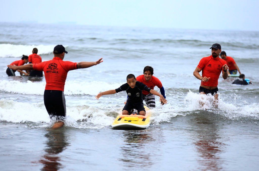 Descrição da imagem #PraTodosVerem: Imagem no formato retangular, na horizontal. Um menino está no mar, surfando em uma prancha adaptada. O menino usa uma roupa de surfe, preta, e sua prancha é amarela. Alguns homens, de camiseta vermelha, estão acompanhando o menino, de perto, para garantir sua segurança. Fim da descrição.