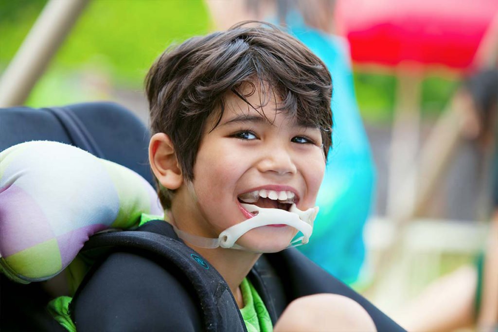 Descrição da imagem #PraTodosVerem: Imagem no formato retangular, na horizontal. Um menino que tem paralisia cerebral está sentado em sua cadeira de rodas. Ele é moreno, tem a pele clara e cabelos castanhos, lisos e curtos. O menino está sorrindo. Fim da descrição.