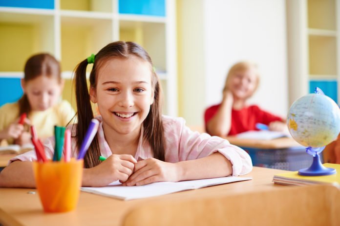 Descrição da imagem #PraTodosVerem: A imagem está no formato retangular, na horizontal. Três crianças estão na sala da aula. Em destaque vemos uma menina sorrindo. Atrás dela estão outra menina e um menino. Fim da descrição.