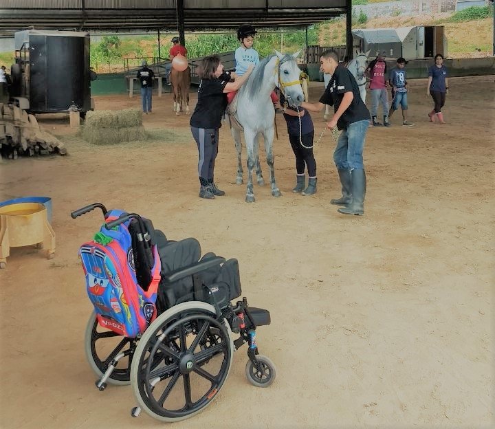 Descrição da imagem #pracegover: O setting de equoterapia. Vemos, em primeiro plano, uma cadeira de rodas. Em segundo plano, vemos uma criança, montada no cavalo, durante a sessão de equoterapia. Fim da descrição.