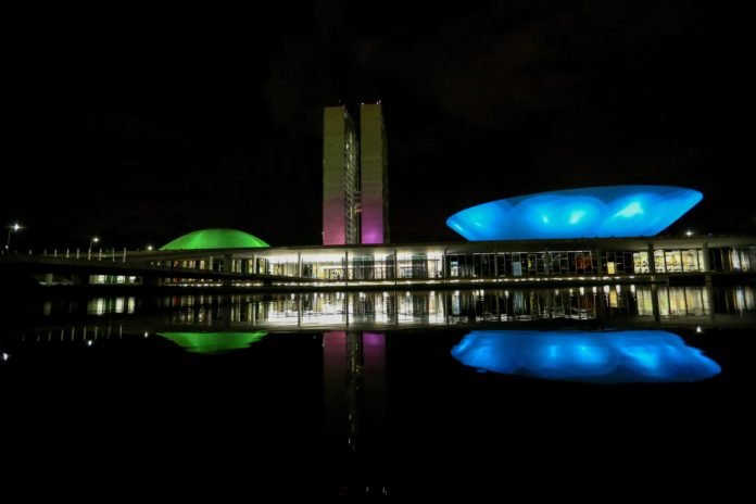 Descrição da imagem #PraCegoVer: Imagem no formato retangular, na horizontal. O Congresso Nacional, durante a noite, iluminado com as cores verde, rosa, azul e roxo, em homenagem ao Dia Mundial das Doenças Raras. Fim da descrição.