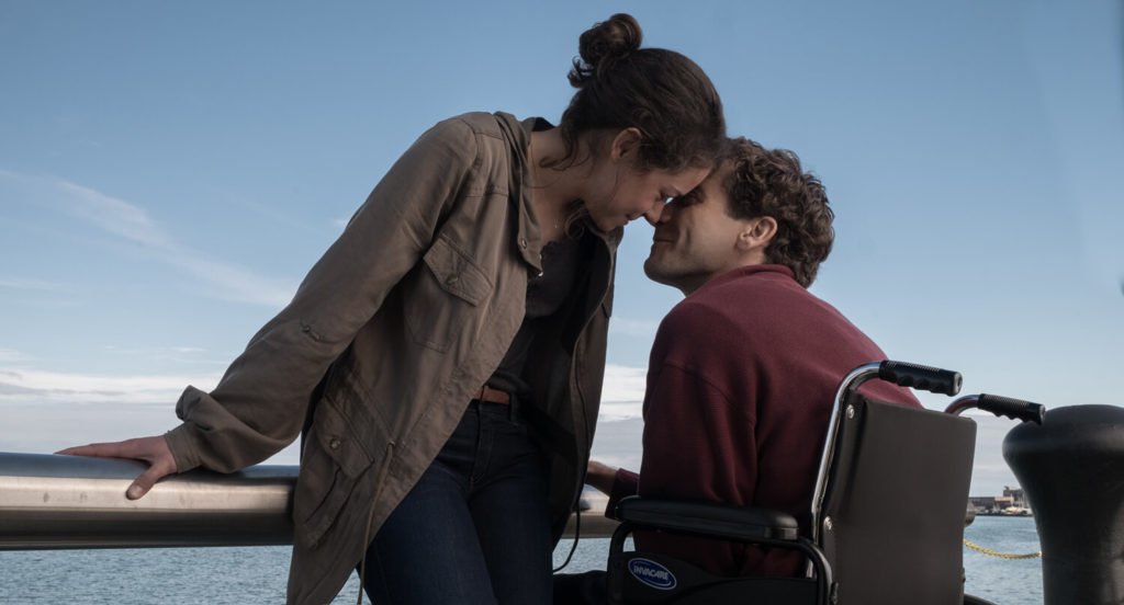 Descrição da imagem #PraCegoVer: Imagem no formato retangular, na horizontal. Cena do filme O que te faz mais forte. Os protagonistas estão se beijando. A mulher está de pé e o rapaz está sentado em sua cadeira de rodas. Fim da descrição.