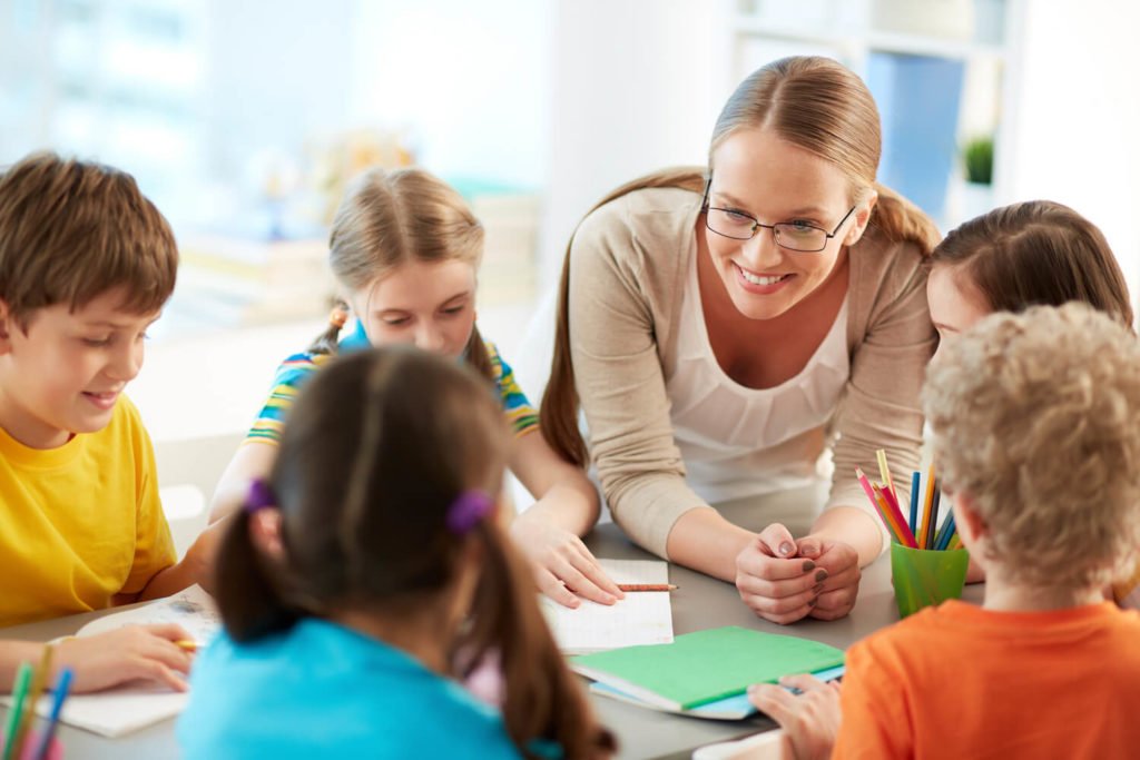 Descrição da imagem #PraTodosVerem: A imagem está no formato retangular, na horizontal. Um grupo de alunos em uma sala de aula. Eles estão sentados em uma mesa, e a professora está entre eles, os orientando sobre uma atividade. Fim da descrição.