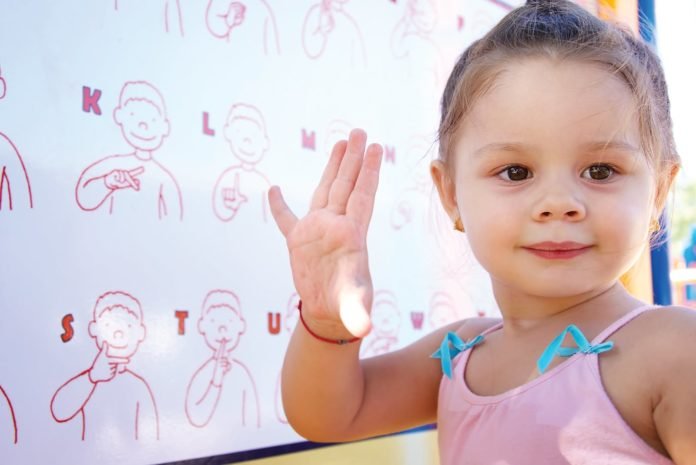 Descrição da imagem: foto de uma menina. Ela está com a mão direita levantada para fazer um sinal em Libras. Atrás dela vemos um painel com os sinais em Libras. Fim da descrição.
