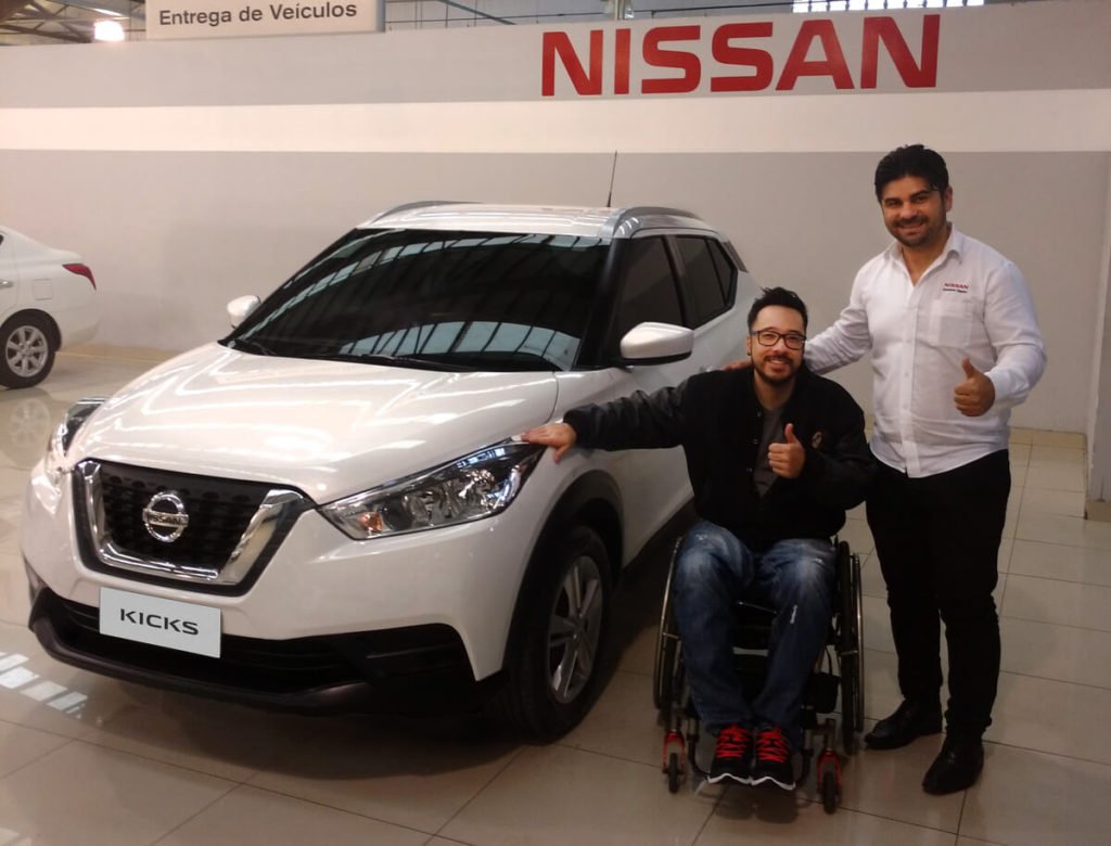 Descrição da imagem #PraCegoVer: Imagem no formato retangular, na horizontal. O primeiro comprador de um Kicks S Direct, no Brasil, e o vendedor. Eles estão ao lado de um Kicks, branco. Ao fundo está o logo da Nissan, em letras vermelhas. O comprador está em sua cadeira de rodas e o vendedor está de pé. Fim da descrição.