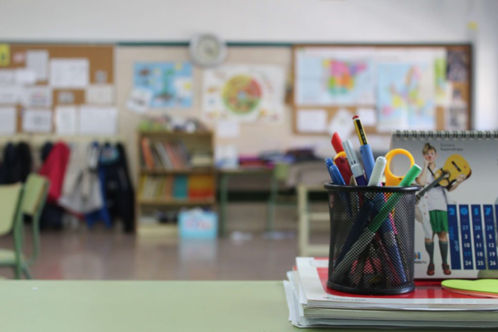 Descrição da imagem #PraCegoVer: A imagem está no formato retangular, na horizontal. Uma sala de aula, vista da mesa do professor. Em cima da mesa estão objetos como porta-lápis e livros. Ao fundo vemos uma parede com vários trabalhos dos alunos, expostos. Fim da descrição.