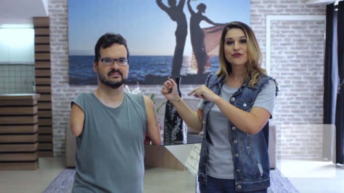 Descrição da imagem #PraCegoVer: Imagem na retangular, na vertical. Dudé e Renata durante a gravação do programa. Ao fundo vemos uma imagem da silhueta de um casal, na praia. Em primeiro plano, temos Dudé, que usa óculos, é moreno e tem pele clara, cabelo e barba castanhos. Dudé não tem os braços. Ele usa uma regata azul. Renata, que está ao seu lado, tem pele clara, cabelos longos e loiros. Ela usa uma blusa cinza e um colete jeans. Fim da descrição.