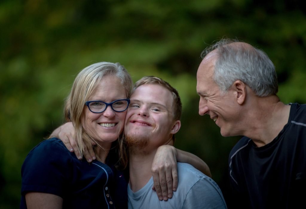 Descrição da imagem #PraCegoVer: Um jovem, com síndrome de Down, está acompanhado de seus pais. Eles estão abraçados. O jovem está no meio do casal. Todos estão sorrindo. Ao fundo, vemos uma área verde. Fim da descrição.
