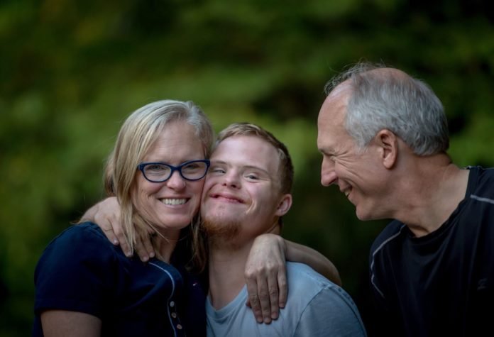Descrição da imagem #PraCegoVer: Um jovem, com síndrome de Down, está acompanhado de seus pais. Eles estão abraçados. O jovem está no meio do casal. Todos estão sorrindo. Ao fundo, vemos uma área verde. Fim da descrição.