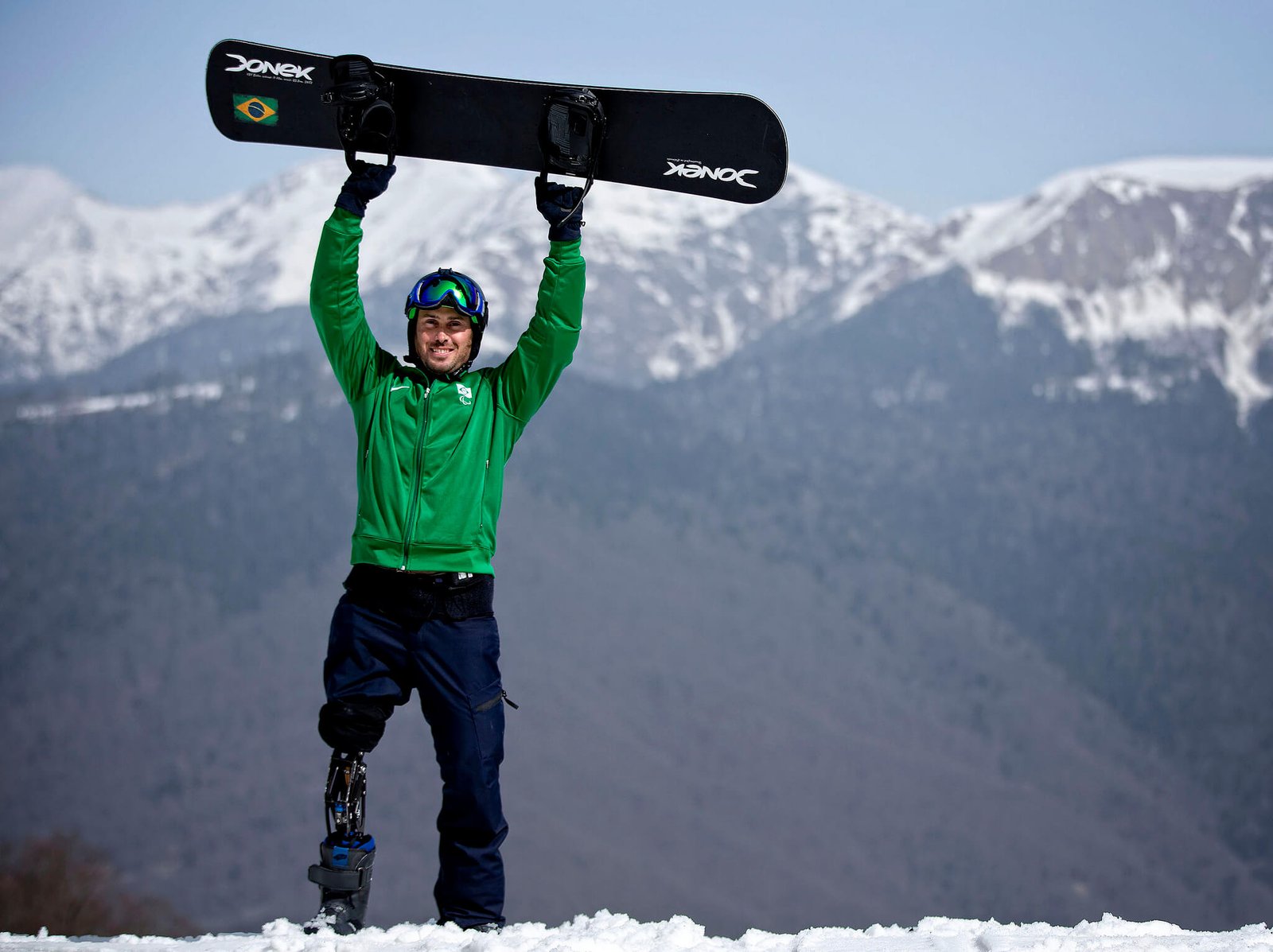 Descrição da imagem #PraCegoVer: O paratleta André Cintra. Ele usa uma prótese na perna direita e segura seu snowboard. Ao fundo, vemos montanhas cobertas de neve. Fim da descrição.