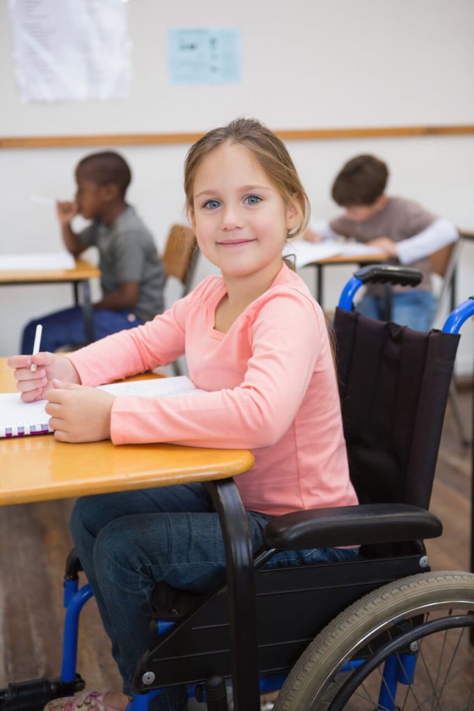 Descrição da imagem #PraCegoVer: Imagem no formato retangular, na vertical. Uma menina cadeirante na sala de aula. Ela está em primeiro plano. Ao fundo, vemos dois estudantes sentados em suas carteiras. A menina é loira, tem cabelos lisos e compridos. Ela usa uma blusa cor de rosa, de mangas compridas e uma calça jeans, e está sentada em sua cadeira de rodas. A menina olha para a foto e está sorrindo. Fim da descrição.