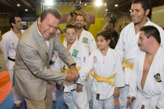Descrição da imagem #PraCegoVer: A imagem está no formato retangular, na horizontal. Arnold Schwarzenegger está comprimentando a equipe de judô formada por atletas com deficiência intelectual. Fim da descrição.