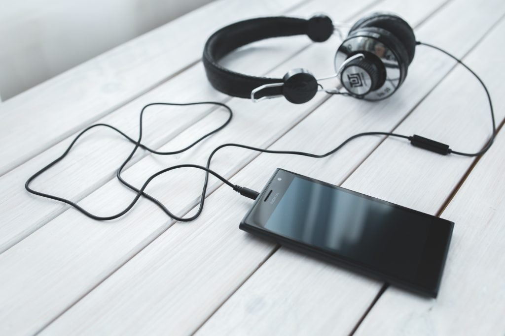 Descrição da imagem #pratodosverem: Um fone de ouvido está conectado a um celular. Eles estão em cima de uma superfície de madeira branca. Fim da descrição.