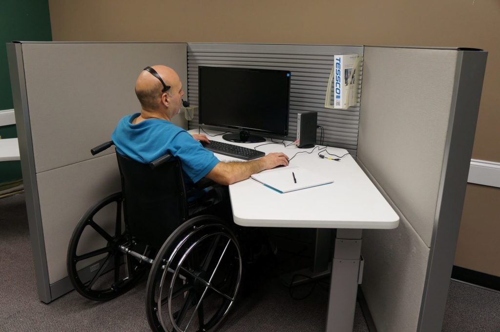 Descrição da imagem #PraCegoVer: Imagem no formato retangular, na horizontal. Um homem cadeirante está trabalhando. Ele está sentado em frente a uma mesa branca, com um computador. Fim da descrição