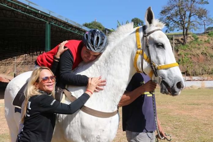 Descrição da imagem #PraCegoVer: Imagem no formato retangular, na horizontal. Um rapaz está deitado no dorso de um cavalo branco. Ao lado do cavalo, dois profissionais, dão apoio ao rapaz. Fim da descrição.
