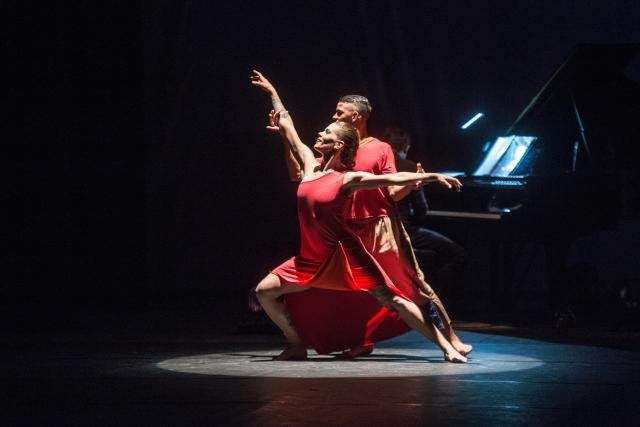Descrição da imagem #PraCegoVer: Imagem no formato retangular, na horizontal. Os bailarinos Roberta e Deivid durante apresentação da Cia. de Dança Lápis de Seda. Eles usam roupas vermelhas e estão em um palco, com fundo preto, fazendo uma coreografia. Fim da descrição.