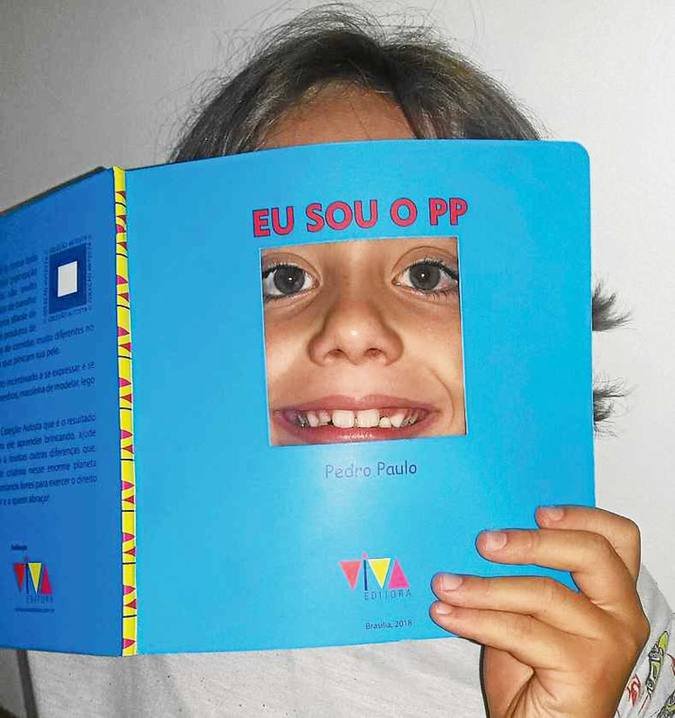 Descrição da imagem #PraCegoVer: Imagem no formato quadrado. O menino está com o rosto na capa do livro. O livro tem capa azul e uma abertura, onde é possível colocar o rosto. Ele está sorrindo. Fim da descrição.