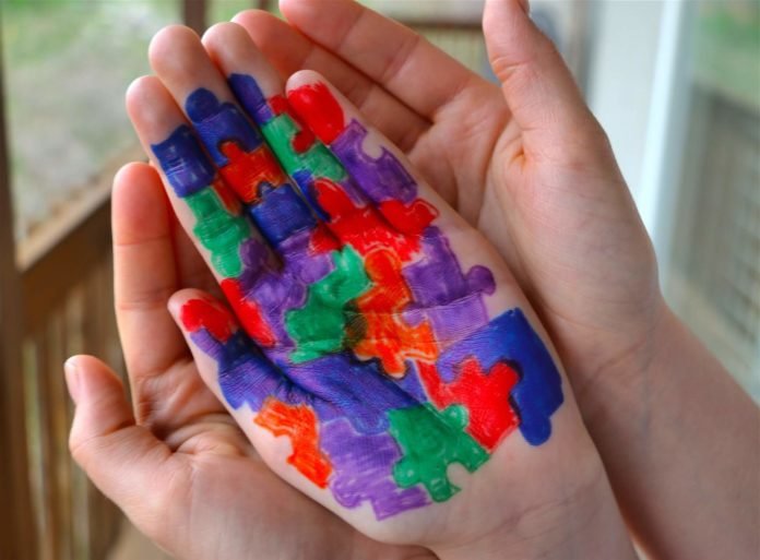 Descrição da imagem #PraCegoVer: Imagem no formato retangular, na horizontal. Duas mãos estão abertas e sobre elas está uma mão pintada com a imagem de peças de um quebra-cabeça, nas cores vermelho, azul, roxo e verde, símbolo do autismo. Fim da descrição.