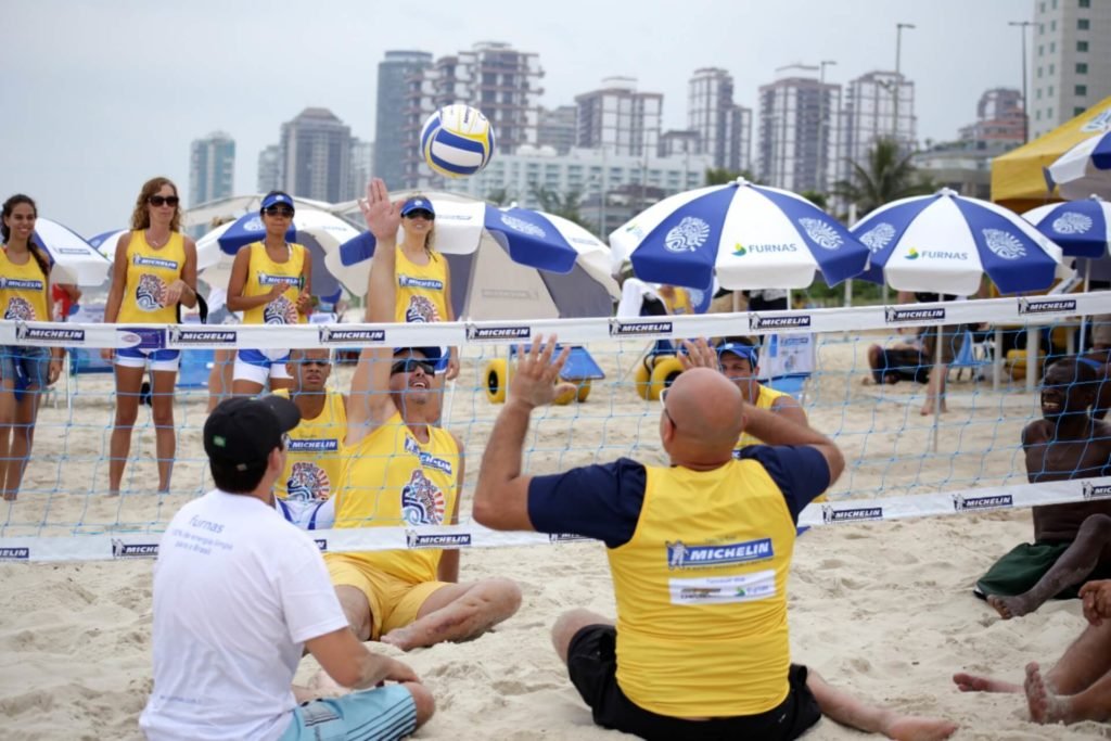 Descrição da imagem #PraCegoVer: Imagem no formato retangular, na horizontal. Um grupo de pessoas jogando vôlei sentado, na praia. Fim da descrição.