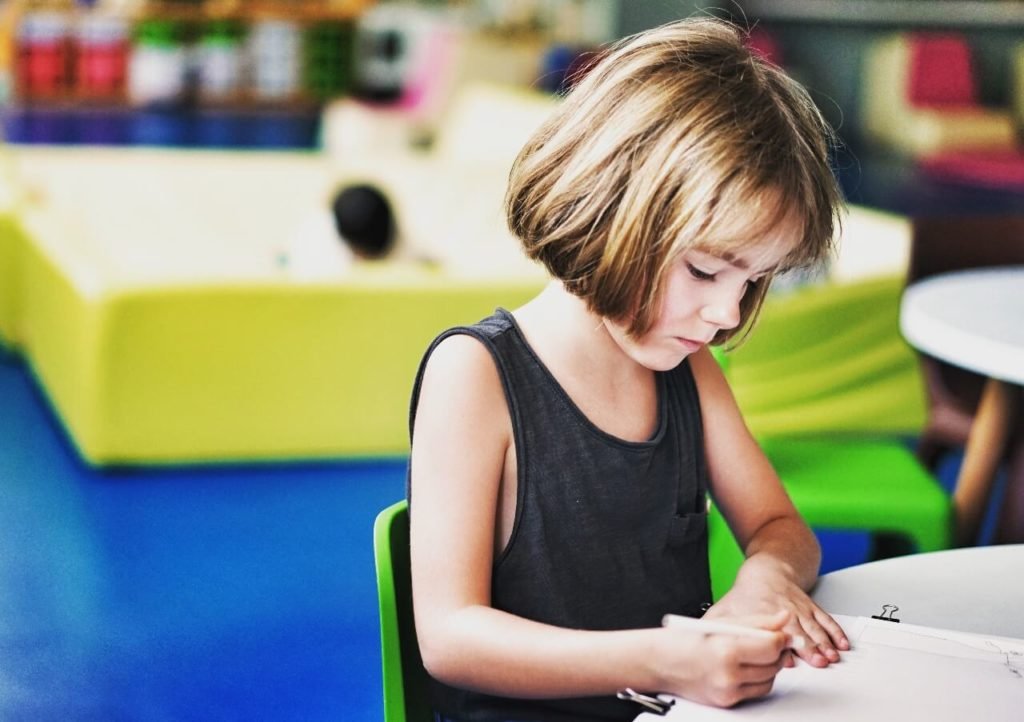 Descrição da imagem #PraCegoVer: Imagem no formato retangular, na horizontal. Uma menina está sentada em uma sala, com objetos coloridos. Ela está fazendo uma atividade em um caderno. Fim da descrição.