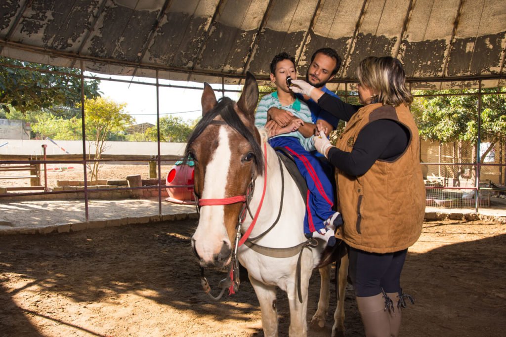 Descrição da imagem #PraCegoVer: Imagem no formato retangular, na horizontal. Um rapaz está montado em um cavalo branco com manchas marrons. Ele está sendo apoiado por um profissional de equoterapia, que também está montado no cavalo. Ao lado do cavalo, a fonoaudióloga Andrea Ribeiro, realiza o atendimento do rapaz. Eles estão no setting terapêutico. Fim da descrição.
