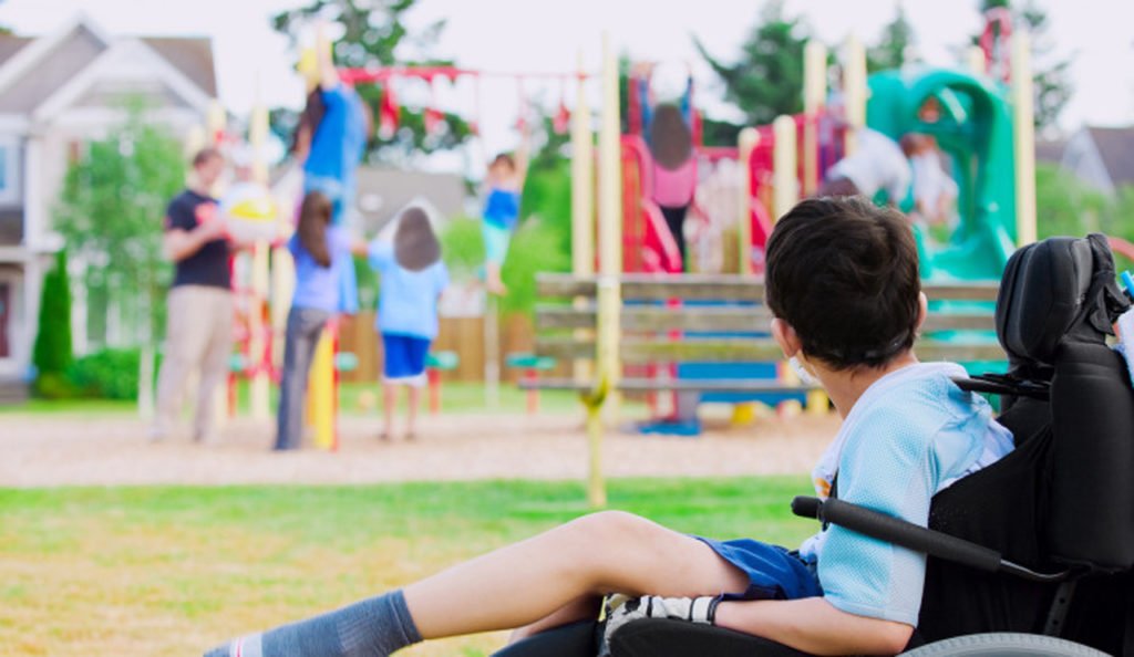 Descrição da imagem #PraCegoVer: Imagem no formato retangular, na horizontal. Um menino está sentado em sua cadeira de rodas. Ele observa as crianças brincando em um parque de diversões. Fim da descrição.
