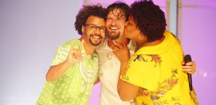 Descrição da imagem #PraCegoVer: Imagem no formato retangular, na horizontal. O cantor Saulo está entre o casal Marcos e Gabriela. Marcos faz o sinal universal de amor na língua de sinais e Gabriela dá um beijo no cantor, que sorri. Fim da descrição.