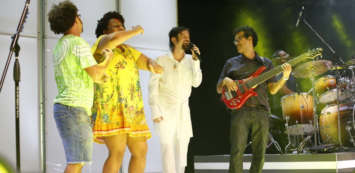 Descrição da imagem #PraCegoVer: Imagem no formato retangular, na horizontal. Palco do show do cantor Saulo. Ao seu lado esquerdo estão o casal Marcos e Gabriela e do seu lado direito está o guitarrista de sua banda. Fim da descrição.