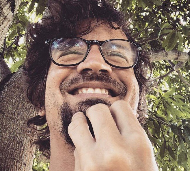 Descrição da imagem #PraCegoVer: Imagem no formato quadrado. Close do rosto do cantor Saulo, ele está fazendo seu sinal em Libras. Saulo está com três dedos no queixo e sorri. Fim da descrição.