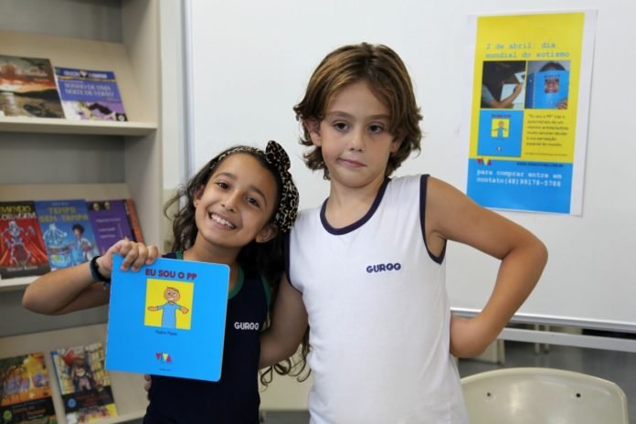Descrição da imagem #PraCegoVer: Imagem no formato retangular, na horizontal. O menino Pedro Paulo está ao lado de uma amiga do colégio, que segura seu livro e sorri. Fim da descrição.