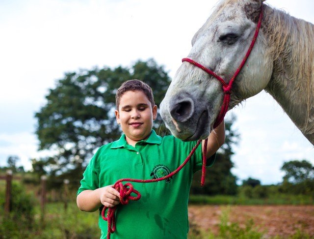 Descrição da imagem #PraCegoVer: Imagem no formato retangular, na horizontal. Gabriel ao lado do cavalo Pé de Pano. Gabriel é um menino moreno, de pele clara. Pé de Pano é um cavalo branco. Fim da descrição.