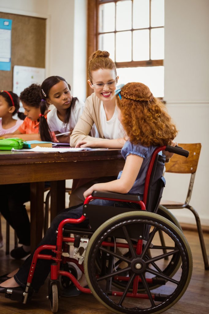 Descrição da imagem #PraCegoVer: Imagem no formato retangular, na vertical. Uma menina, cadeirante, está sentada ao lado de uma mulher. Elas estão em uma sala de aula, ao lado de outros estudantes, que aparecem em segundo plano. Fim da descrição.
