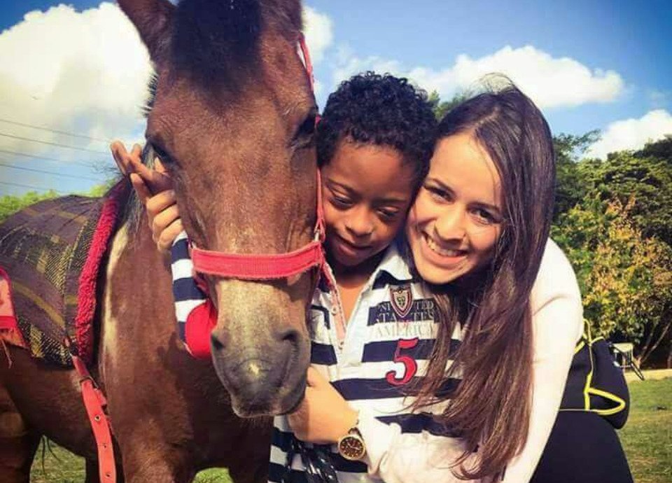 Descrição da imagem: Foto da terapeuta ocupacional Rebecca Martins está abraçando um menino com síndrome de Down. Eles estão ao lado de um cavalo marrom. Fim da descrição.
