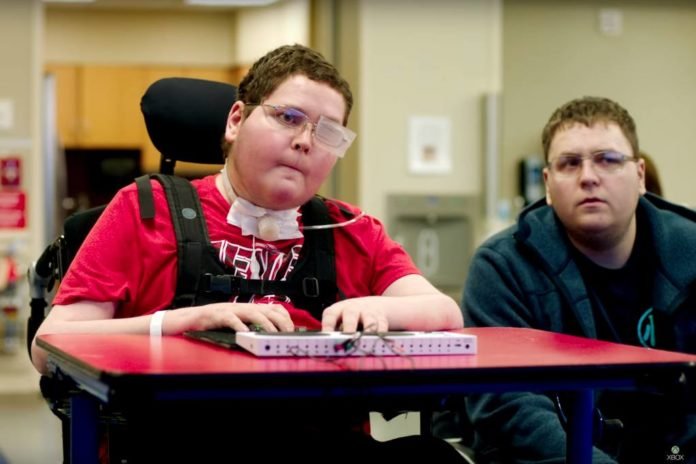 Descrição da imagem #PraCegoVer: Imagem no formato retangular, na horizontal. Foto de um rapaz com paralisia cerebral jogando video game. Ele está sentado em sua cadeira de rodas, com a adaptação de uma mesa e tem o novo controle da Microsoft nas mãos. Ao seu lado está um rapaz, que acompanha seu teste no novo equipamento. Fim da descrição.