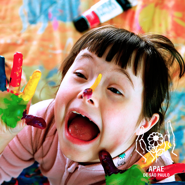 Descrição da imagem #PraCegoVer: Imagem no formato quadrado. Uma menina com síndrome de Down está com as mãos e o nariz pintados. Ela está em meio a muitas tintas, coloridas. Fim da descrição.