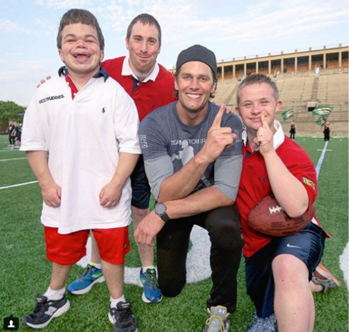 Descrição da imagem #PraCegoVer: Imagem no formato retangular, na horizontal. Três jovens com deficiência intelectual, atendidos pela Best Buddies, estão em um estádio, ao lado do jogador de futebol americano Tom Brady. Eles estão sorrindo. Fim da descrição.