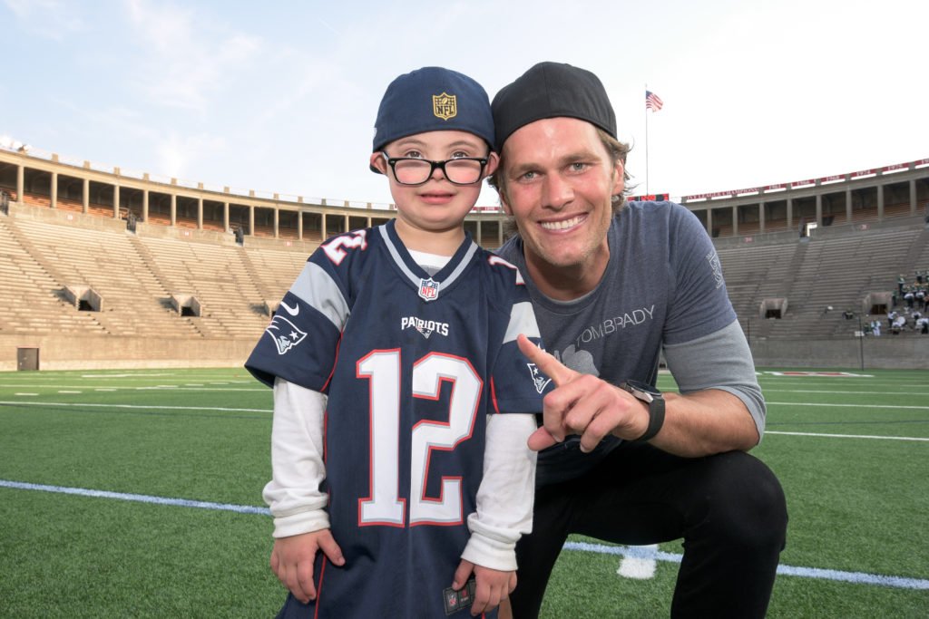 Descrição da imagem #pracegover: Um menino, com síndrome de Down, atendido pela Best Buddies, está em um estádio, ao lado do jogador de futebol americano Tom Brady. Eles estão sorrindo, e o menino está usando uma camisa do time de Brady. Fim da descrição.