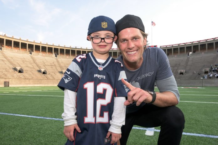 Descrição da imagem #pracegover: Um menino, com síndrome de Down, atendido pela Best Buddies, está em um estádio, ao lado do jogador de futebol americano Tom Brady. Eles estão sorrindo, e o menino está usando uma camisa do time de Brady. Fim da descrição.