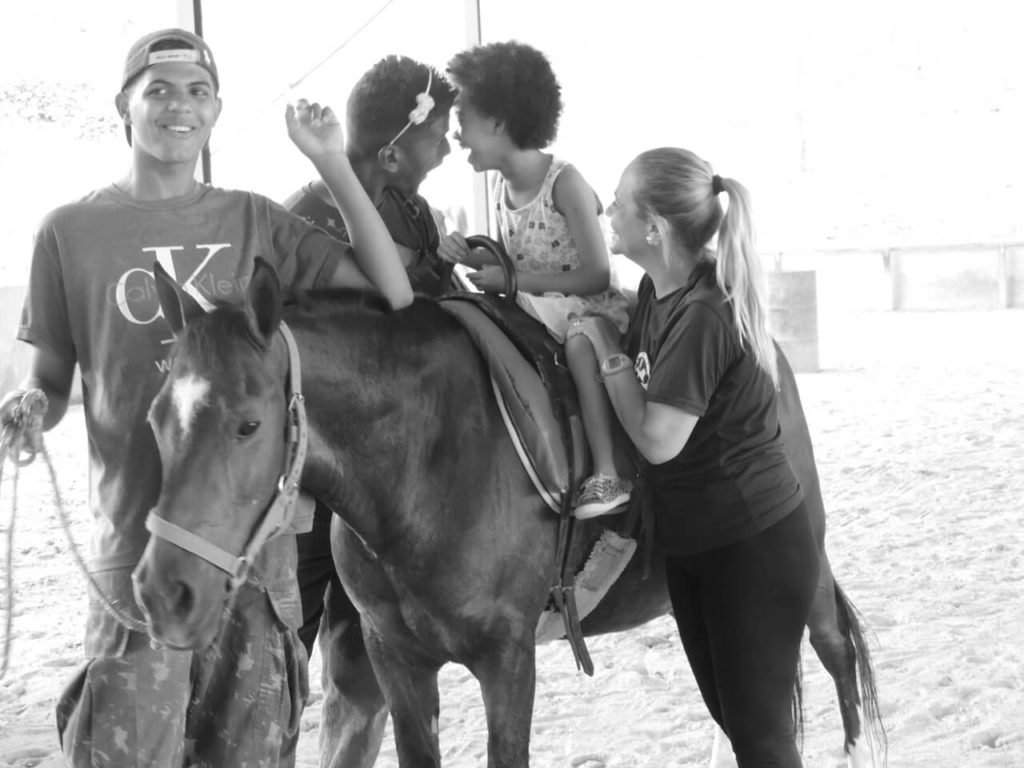 Descrição da imagem: Foto branco e preto de uma criança montada em um cavalo. Ela está sendo apoiada por dois profissionais de equoterapia. Cada um deles está de um lado do cavalo. Eles estão no setting terapêutico. Fim da descrição.