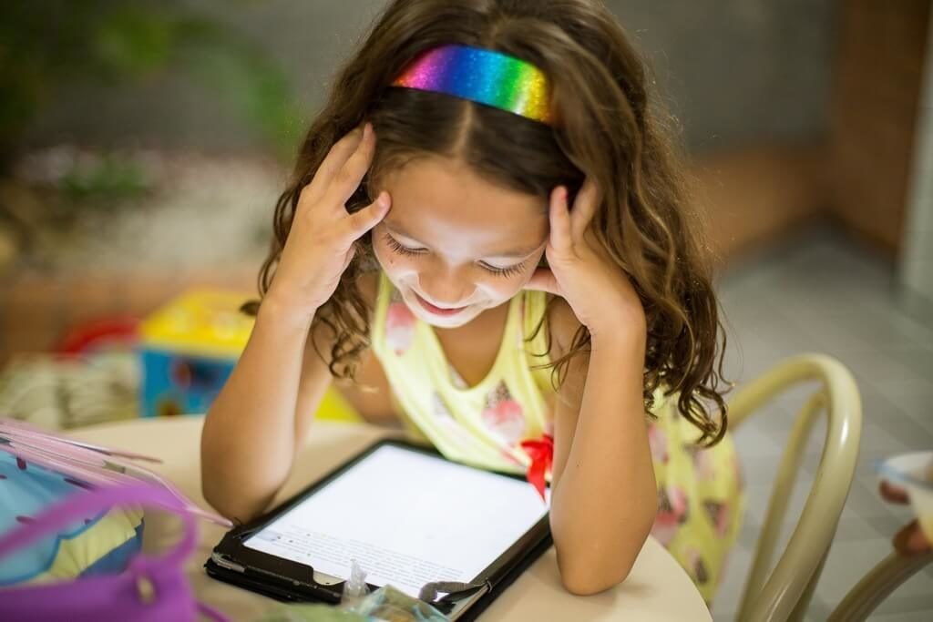 Descrição da imagem #PraCegoVer: Imagem no formato retangular, na horizontal. Uma menina está sentada, vendo um tablet, que está na sua frente. A menina está com as mãos na cabeça. Ela é morena, tem cabelos castanhos, longos. Sobre eles ela usa uma tiara colorida. Fim da descrição.