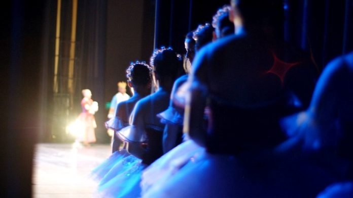 Descrição da imagem: foto de uma fileira de bailarinas à espera do momento em que devem entrar no palco. É possível ver os detalhes do tule de suas saias. Ao fundo, vemos uma bailarina durante apresentação, no palco principal. Fim da descrição.