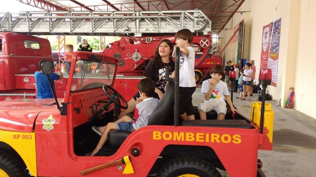 Descrição da imagem #PraCegoVer: Imagem no formato retangular, na horizontal. Várias crianças estão dentro de um jeep dos Bombeiros. O jeep é vermelho e tem detalhes em amarelo. Fim da descrição.