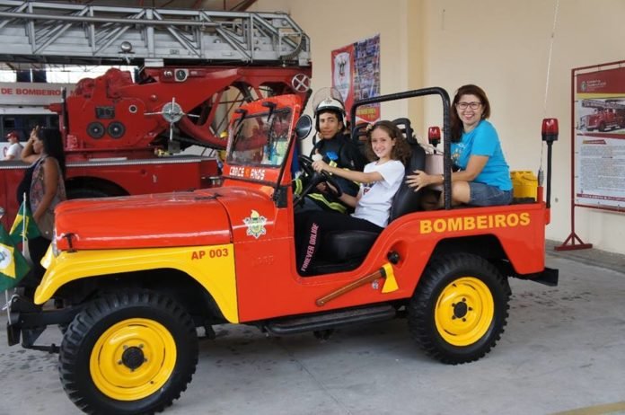 Descrição da imagem #PraCegoVer: Imagem no formato retangular, na horizontal. Três pessoas estão dentro de um jeep dos Bombeiros. No banco da frente estão um bombeiro e uma menina, e no banco traseiro, está uma mulher. O jeep é vermelho e tem detalhes em amarelo. Fim da descrição.