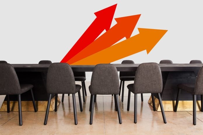 Descrição da imagem #PraCegoVer: Imagem no formato retangular, na horizontal. Uma sala de reuniões, com uma mesa grande e várias cadeiras. Na parede, branca, vemos a ilustração de três setas no sentido ascendente. Fim da descrição.