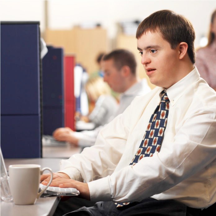 Descrição da imagem #PraCegoVer: Um jovem com síndrome de Down está sentado em uma mesa, em uma empresa. Ele usa uma camisa branca e gravata. Está usando um computador. Fim da descrição.