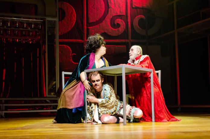 Descrição da imagem #PraCegoVer: Imagem no formato retangular, na horizontal. Cena da montagem teatral Molière. Um homem e uma mulher estão sentados em frente a uma mesa, e em baixo dela, está um homem. Todos os personagens estão caracterizados. Fim da descrição.
