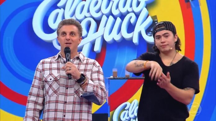 Descrição da imagem #PraCegoVer: Imagem no formato retangular, na horizontal. Imagem do programa Caldeirão do Huck. O apresentador Luciano Huck está ao lado de Whindersson Nunes, que simula sinais de Libras. Fim da descrição.